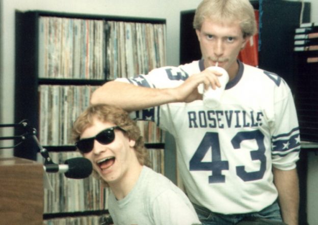 Chris Kelly in his Risky Business glasses talking into the mic and Jon "Stix" Monroe with his elbow on Chris' head of the WHEN Rock Machine show circa 1988. LPs are in shelves behind them.